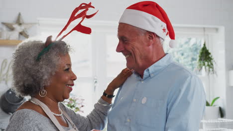 Liebevolles-älteres-Paar-Mit-Kostümgeweihen-Tanzt-In-Der-Küche,-Während-Es-Das-Weihnachtsessen-Zubereitet