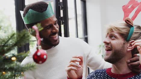 Man-Putting-Fancy-Dress-Antlers-On-Male-Partner-As-They-Hang-Decorations-On-Christmas-Tree-Together