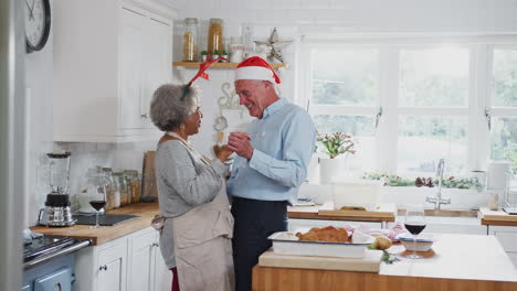 Loving-Senior-Couple-Wearing-Fancy-Dress-Antlers-Dance-In-Kitchen-Whilst-Preparing-Christmas-Dinner