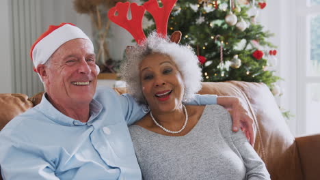 Portrait-Of-Senior-Couple-Wearing-Reindeer-Antlers-And-Santa-Hat-Sitting-On-Sofa-On-Christmas-Day