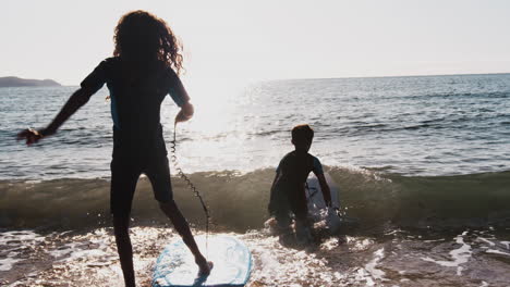 Zwei-Silhouettierte-Kinder-In-Neoprenanzügen-Spielen-Im-Sommerurlaub-Am-Strand-Mit-Bodyboards-Im-Meer