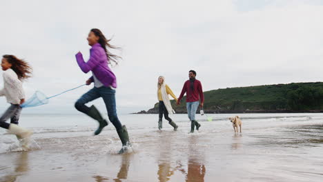 Multi-Cultural-Family-With-Pet-Dog-Walking-Along-Beach-Shoreline-On-Winter-Vacation