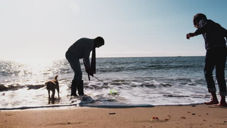 Silhouette-Von-Vater-Und-Sohn,-Die-Mit-Netz-Und-Hund-An-Brechenden-Wellen-Am-Strand-Spielen