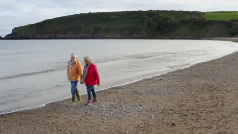 Drone-Shot-Of-Senior-Couple-Holding-Hands-As-They-Walk-Along-Shoreline-On-Winter-Beach-Vacation