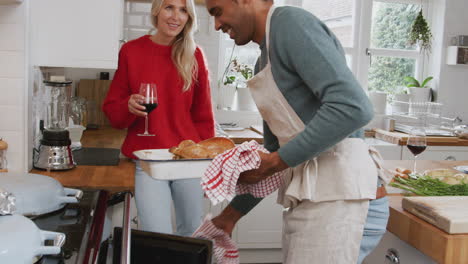 Man-Taking-Turkey-Out-Of-Oven-As-Couple-Drink-Wine-And-Prepare-Dinner-On-Christmas-Day