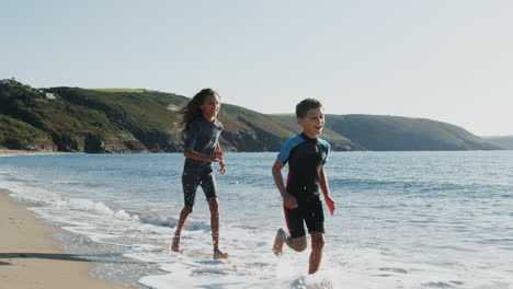 Zwei-Kinder-In-Neoprenanzügen-Laufen-Im-Sommerurlaub-Am-Strand-Durch-Die-Wellen