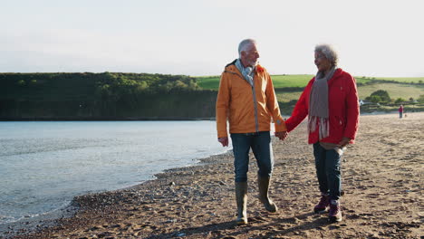 Loving-Active-Senior-Couple-Holding-Hands-As-They-Walk-Along-Shoreline-On-Winter-Beach-Vacation