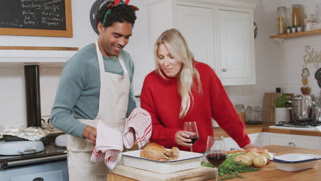Mann-Holt-Truthahn-Aus-Dem-Ofen,-Während-Ein-Paar-Am-Weihnachtstag-Wein-Trinkt-Und-Das-Abendessen-Zubereitet