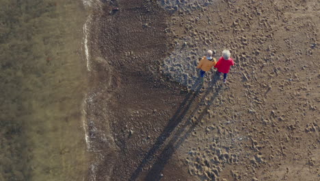 Drone-Shot-Of-Senior-Couple-Holding-Hands-As-They-Walk-Along-Shoreline-On-Winter-Beach-Vacation