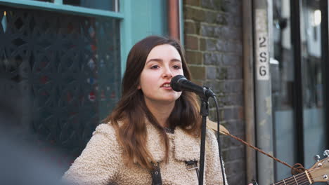 Female-Musician-Busking-Playing-Acoustic-Guitar-And-Singing-To-Crowd-Outdoors-In-Street
