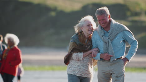 Loving-Active-Senior-Couple-Walk-Arm-In-Arm-Along-Shoreline-On-Winter-Beach-Vacation
