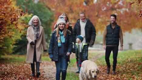 Smiling-Multi-Generation-Family-With-Dog-Walking-Along-Path-Through-Autumn-Countryside-Together