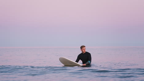 Man-Wearing-Wetsuit-Sitting-And-Floating-On-Surfboard-At-Sea-As-Waves-Break-Around-Him