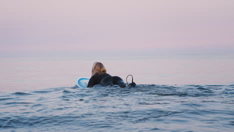 Rear-View-Of-Woman-Wearing-Wetsuit-Paddling-Surfboard-Out-To-Sea