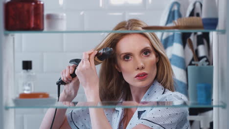 View-Through-Bathroom-Cabinet-Of-Young-Woman-Wearing-Pajamas-Curling-Hair
