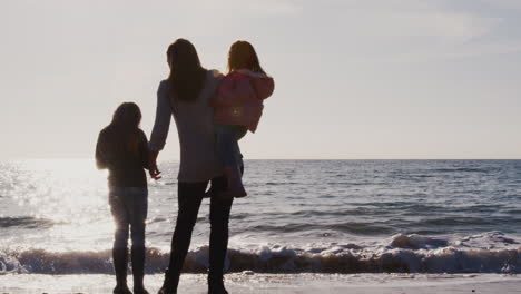 Rear-View-Of-Mother-With-Daughters-Looking-Out-To-Sea-Silhouetted-Against-Sun