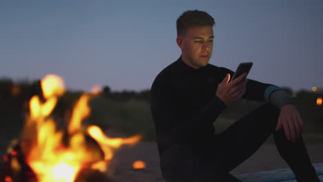 Man-Sitting-On-Surfboard-By-Camp-Fire-On-Beach-Using-Mobile-Phone-As-Sun-Sets-Behind-Her