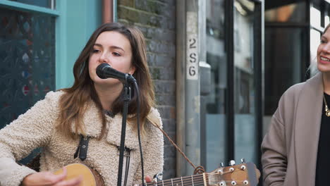 Female-Musician-Busking-Playing-Acoustic-Guitar-And-Singing-To-Crowd-Outdoors-In-Street