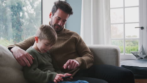 Father-Playing-Video-Game-With-Son-On-Mobile-Phone-Sitting-On-Sofa-At-Home