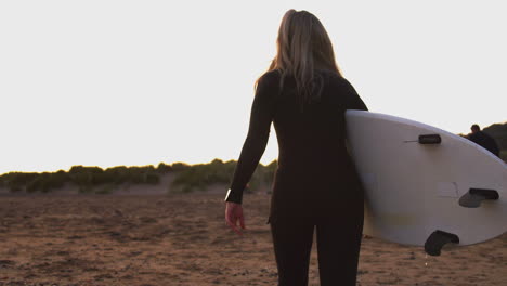 Rear-View-Of-Woman-Wearing-Wetsuit-Carrying-Surfboard-Up-Beach-After-Surfing