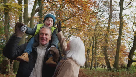 Grandparents-Walking-With-Grandson-On-Grandfathers-Shoulders-Along-Path-In-Autumn-Countryside