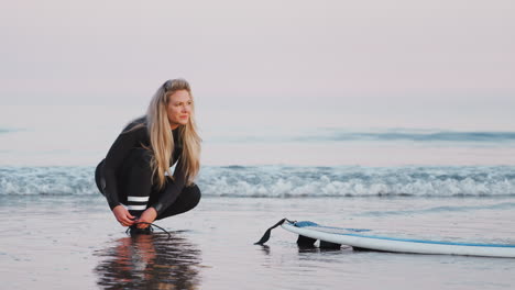Female-Surfer-Wearing-Wetsuit-Attaching-Surfboard-Leash-To-Ankle-In-Waves