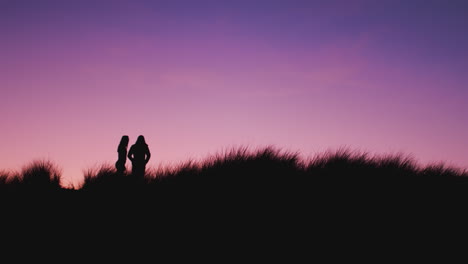 Silhouette-Of-Couple-At-Beach-Standing-On-Sand-Dunes-Covered-In-Grass-Against-Setting-Sun