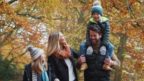 Lächelnde-Familie-Geht-Den-Weg-Durch-Die-Herbstliche-Landschaft-Entlang,-Während-Der-Vater-Seinen-Sohn-Auf-Den-Schultern-Trägt