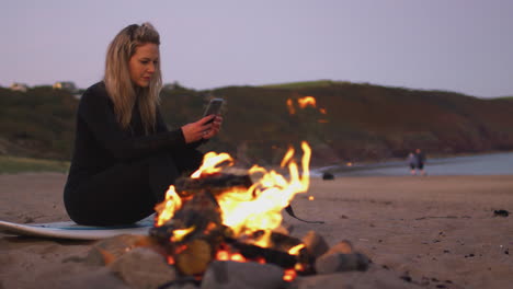 Woman-Sitting-On-Surfboard-By-Camp-Fire-On-Beach-Using-Mobile-Phone-As-Sun-Sets-Behind-Her