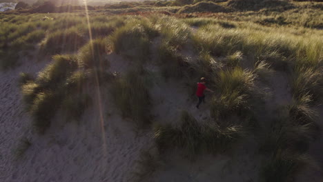 Drohnenaufnahme-Eines-Kleinen-Jungen-Im-Strandurlaub,-Der-In-Sanddünen-Gegen-Die-Flackernde-Sonne-Spielt