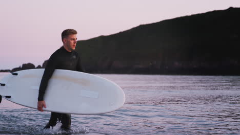 Man-Wearing-Wetsuit-Carrying-Surfboard-As-He-Walks-Out-Of-Sea