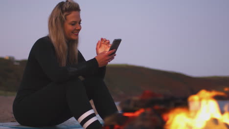 Woman-Sitting-On-Surfboard-By-Camp-Fire-On-Beach-Using-Mobile-Phone-As-Sun-Sets-Behind-Her