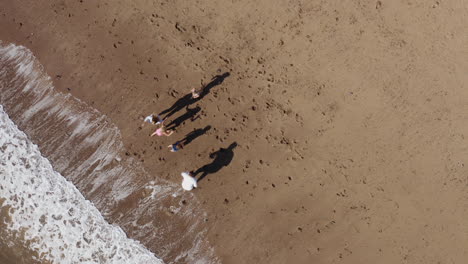 Toma-De-Drone-De-Una-Familia-De-Vacaciones-Corriendo-Por-La-Playa-Hacia-Las-Olas-Rompientes