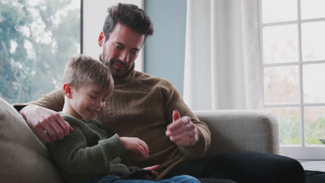 Father-Playing-Video-Game-With-Son-On-Mobile-Phone-Sitting-On-Sofa-At-Home