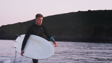 Man-Wearing-Wetsuit-Carrying-Surfboard-As-He-Walks-Out-Of-Sea