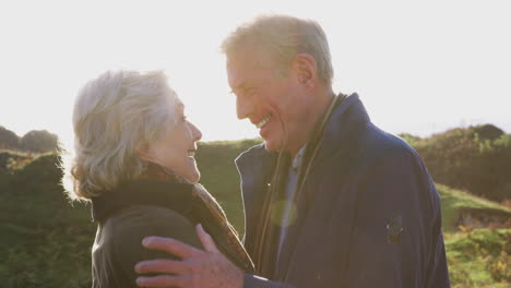 Loving-Active-Senior-Couple-Hugging-As-They-Walk-Along-Coastal-Path-Against-Flaring-Sun