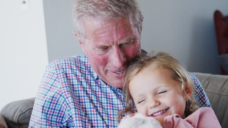 Loving-Grandfather-Cuddling-Granddaughter-Holding-Soft-Toy-Rabbit-At-Home