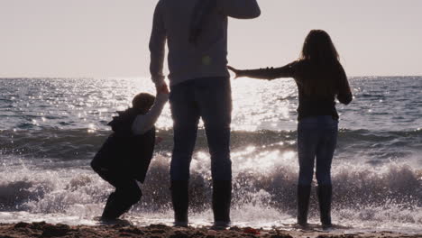 Rückansicht-Eines-Vaters-Mit-Kindern,-Der-über-Wellen-Springt-Und-Auf-Das-Meer-Blickt,-Dessen-Silhouette-Sich-Vor-Der-Sonne-Abzeichnet