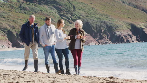 Senior-Couple-Walking-Along-Shoreline-With-Adult-Offspring-On-Winter-Beach-Vacation