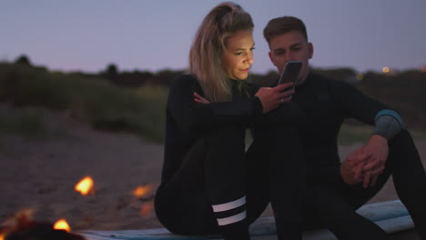 Couple-Sitting-On-Surfboard-By-Camp-Fire-On-Beach-Using-Mobile-Phone-As-Sun-Sets-Behind-Them