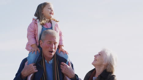 Grandfather-Giving-Granddaughter-Ride-On-Shoulders-As-They-Walk-In-Countryside-With-Grandmother