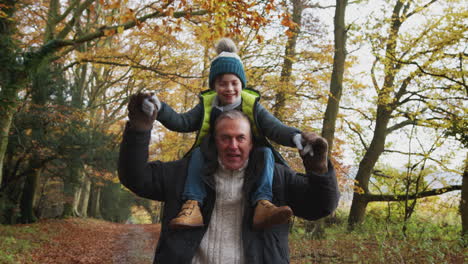 Smiling-Grandfather-Carrying-Grandson-On-Shoulders-As-They-Walk-Along-Path-In-Autumn-Countryside