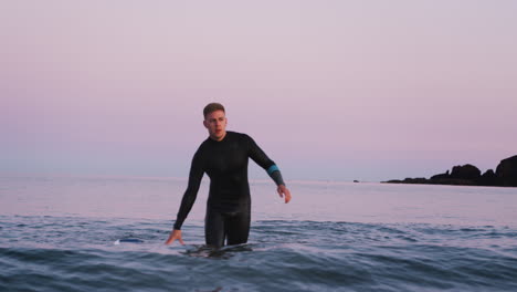 Man-Wearing-Wetsuit-Holding-Floating-Surfboard-As-He-Walks-Through-Waves