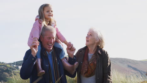 Grandfather-Giving-Granddaughter-Ride-On-Shoulders-As-They-Walk-In-Countryside-With-Grandmother