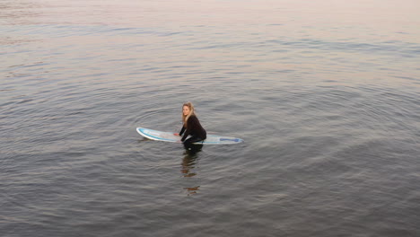 Drone-Shot-Of-Woman-Wearing-Wetsuit-Sitting-On-Surfboard-At-Sea