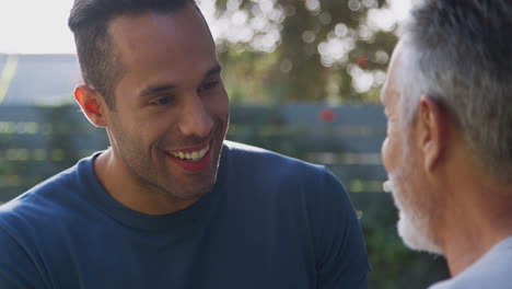 Senior-Hispanic-Man-Talking-And-Laughing-With-Adult-Son-In-Garden-At-Home