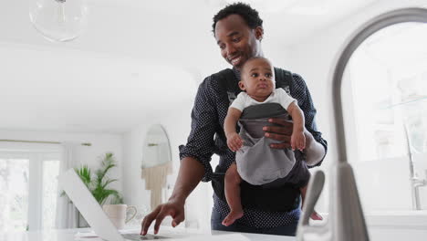 Father-With-Baby-Son-In-Sling-Multi-tasking-Working-From-Home-On-Laptop