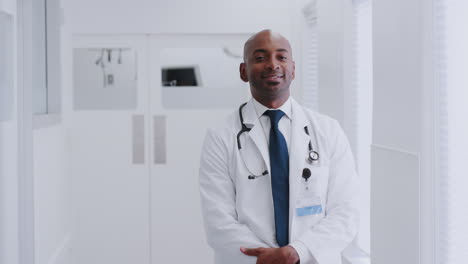 Portrait-Of-Smiling-Mature-Male-Doctor-Wearing-White-Coat-Standing-In-Hospital-Corridor