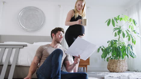 Junges-Paar-Mit-Schwangerer-Frau-Im-Schlafzimmer-Schaut-Sich-Anleitung-Zum-Selbstaufbau-Eines-Babybetts-An