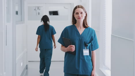 Portrait-Of-Smiling-Female-Doctor-Wearing-Scrubs-In-Busy-Hospital-Corridor-Holding-Digital-Tablet